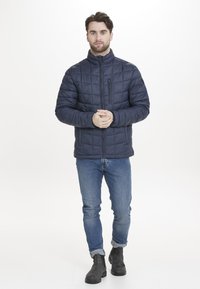 Mörkblå quiltad puffjacka med hög krage, dragkedja fram och sidofickor, bärs över blå jeans och mörka ankelstövlar.