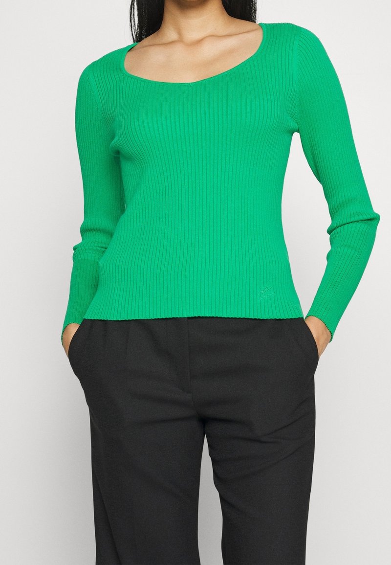 Femme portant un pull vert côtelé ajusté à manches longues avec un col carré et un pantalon noir, mains dans les poches.