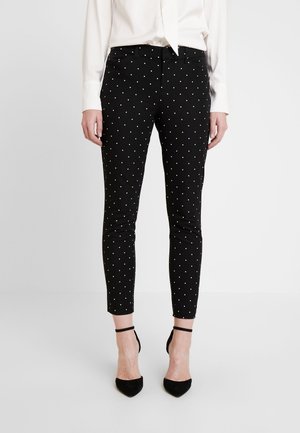 Femme portant un pantalon noir slim à pois blancs, assorti à des talons noirs avec bride à la cheville et un chemisier blanc rentré.