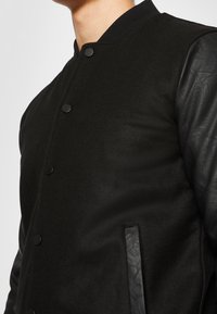 Black varsity jacket featuring wool body and black leather sleeves, with snap buttons and side pockets, smooth texture, and fitted design.