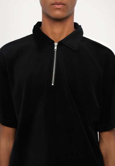 Polo shirt noir à manches courtes en tissu doux, avec un col zippé, des lignes épurées et sans motifs ni accents visibles.