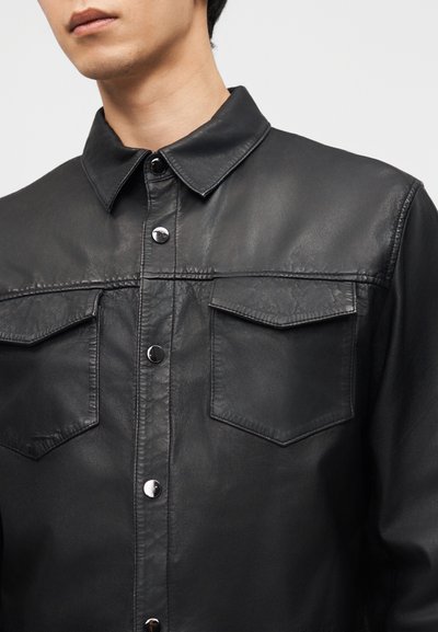 Άνδρας που φοράει μαύρο δερμάτινο πουκάμισο με κουμπιά και θήκες με καπάκι στο στήθος, μπροστά σε απλό ανοιχτό φόντο.