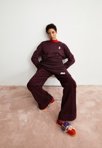 Bordeaux langemouwen shirt met een wit logo, gecombineerd met bijpassende bordeaux corduroy broek. Schoeisel met een rood ontwerp en kleurrijke veters.