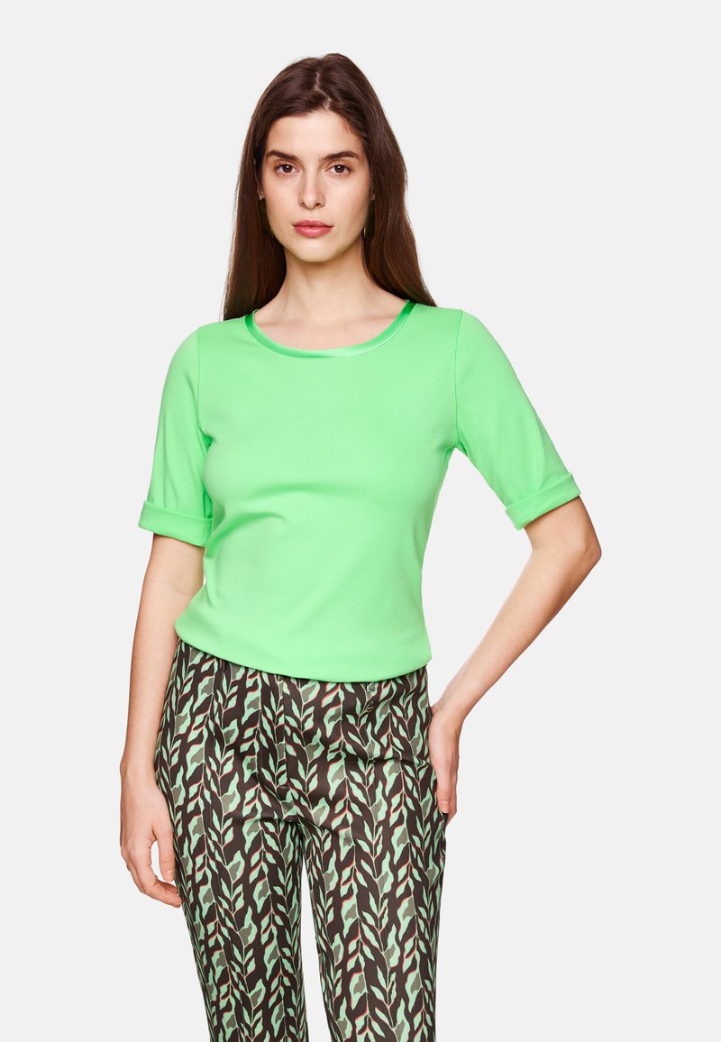 Short-sleeve, round-neck shirt in bright green, made of smooth fabric. Paired with patterned trousers featuring leaf designs in green and brown.