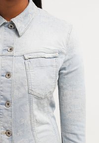 Giacca di jeans azzurro chiaro con un sottile motivo floreale, bottoni metallici, colletto e una singola tasca sul petto con bordo sfilacciato.