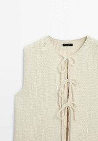 Gilet en tricot crème sans manches avec motif texturé et trois attaches nouées à l'avant, avec un col rond et une étiquette de marque noire à l'intérieur.