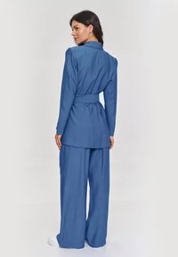 Niebieska marynarka o dopasowanym kroju z paskiem w talii, zestawiona z szerokimi spodniami, wykonana z gładkiego materiału i o zdefiniowanej strukturze.