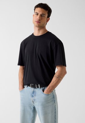 Jonge man met kort krullend haar, gekleed in een zwart T-shirt en lichtblauwe spijkerbroek, staand met handen in de zakken tegen een effen achtergrond.