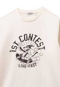 Felpa color crema realizzata in tessuto morbido, con una grafica di un cane dei cartoni animati su un monopattino e il testo nero che recita "1ST CONTEST LIVE FAST."