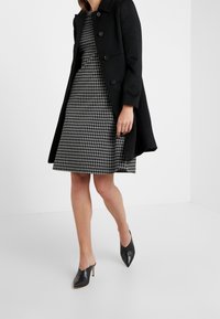 Černý vlněný kabát s knoflíky, nošený přes černo-bílou kostkovanou šaty. Špičaté černé podpatky dotvářejí celý outfit.