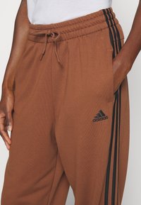 Close-up of brown Adidas sweatpants with black three-stripe detailing on the side, elastic waistband, drawstring, and a hand in the pocket.