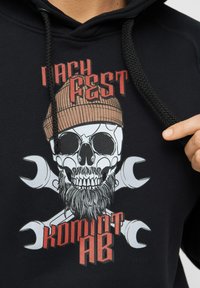 Schwarzer Hoodie mit einem Grafikdesign, das einen Skull mit Bart, gekreuzte Schraubenschlüssel und den Text "I ACH FEST KOMMT AB" in rot und weiß zeigt.