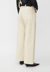Persona che indossa pantaloni larghi di colore avorio e scarpe nere con tacco basso a punta, in piedi con le mani in tasca su uno sfondo neutro.