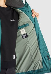 Personne tenant une veste bleu sarcelle ouverte montrant la doublure gris clair et les étiquettes de la marque à l'intérieur, portant un t-shirt noir Khujo en dessous.