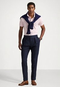 Πόλο πουκάμισο σε ανοιχτό ροζ, πλεκτό μπλε σκούρο πουλόβερ περασμένο στους ώμους, μπλε σκούρο παντελόνι με ρίγες, καφέ δερμάτινα παπούτσια, ασημένιο ρολόι στον καρπό.