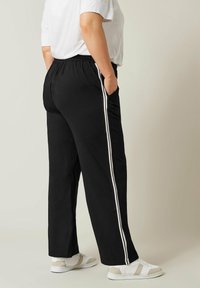 Pantaloni neri con vita elastica e bande laterali bianche, vestibilità leggermente ampia, design semplice. Indossati con scarpe bianche e beige chiaro.