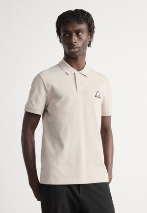 Beige polo skjorte med klassisk krage og korte ermer, med et lite trekantet logo-patch i blått og gult på venstre bryst.
