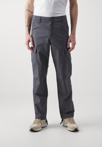 Mann trägt dunkelgraue Cargo-Hosen mit Klapptaschen und beigen Sneakers, steht vor einem schlichten hellen Hintergrund.