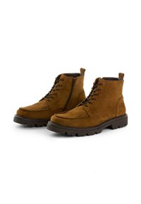 Braune Wildleder-Ankle-Boots mit schwarzen dicken Profilsohlen, Schnürsenkeln und seitlichen Reißverschlüssen, vor einem weißen Hintergrund präsentiert.