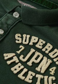 Groene poloshirt met een gebreide textuur kraag, voorzien van crèmekleurige geborduurde tekst: "SUPERB" en "ATHLETICS 7 JPN 4." Twee knoopsluitingen.
