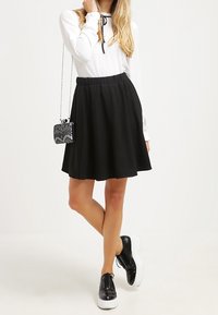 Žena v bílém dlouhém rukávu, černé áčkové sukni, černých platformových botách a s malou vzorovanou taškou přes rameno.