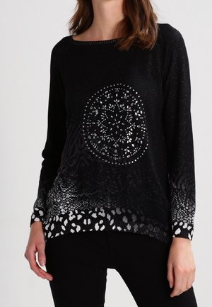 Kvinde iført en sort langærmet sweater med sølvnitter i cirkulært mønster og dyreprintdetaljer på manchetter og kant.