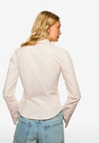 Light pink fitted blouse with a pointed collar, long sleeves, and buttoned cuffs. Smooth fabric with subtle sheen. Worn with high-waisted denim.