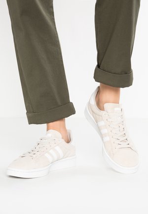 Voeten met beige Adidas sneakers met witte strepen, gecombineerd met opgerolde olijfgroene broeken tegen een egale witte achtergrond.