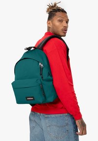 Mochila verde azulado con doble cremallera y bolsillo frontal. Tejido texturizado, correas acolchadas y etiqueta de logo. Llevada por una persona con un sudadera roja.