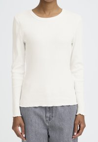 Persona ar pieguļošu baltu ribotu garām piedurknēm, ar salātu malu aprocēm un apakšdaļu, apvienots ar augstām pelēkām džinsu biksēm.