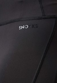 Vêtements de sport noirs arborant le nom de la marque "SNOCKS" en lettres argentées, avec une texture lisse et des détails de couture visibles.