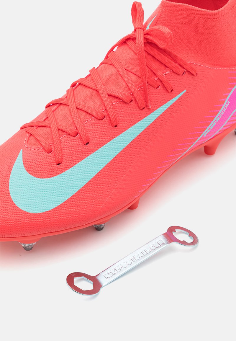 Chaussure de football rose vif avec tige texturée, logo Nike turquoise, six crampons et un outil métallique en forme de clé à molette à côté.