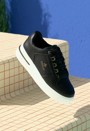 Sneaker in pelle nera con suola bianca e logo "REPLAY", esposto su gradini in piastrelle beige contro uno sfondo di muro blu.