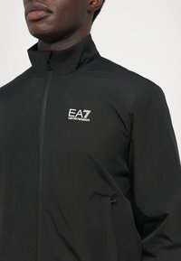 Black zip-up jacket with a high collar, featuring a white EA7 Emporio Armani logo on the upper left side and smooth, lightweight fabric.