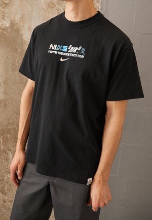 Personne portant un t-shirt noir avec le texte Nike multicolore et le slogan « Un meilleur demain commence aujourd'hui » debout contre un mur texturé.