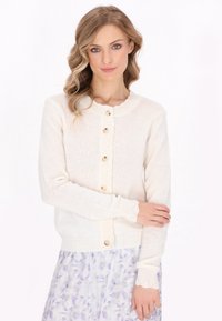 Cardigan crem cu guler dantelat și închidere cu nasturi. Textură tricotată, mâneci lungi cu manșete dantelate, asortat cu o fustă florală.