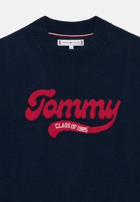 Marinblå stickad tröja med en stor röd "Tommy"-logotyp och texten "CLASS OF 1985", med ribbad rund halsringning och detaljer vid fållen.