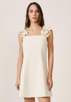 Mujer con vestido crema sin mangas, escote cuadrado y adornos florales en 3D en las tiras de los hombros, posando frente a un fondo liso.