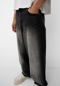 Široké černé džíny vyrobené z denimu s vybledlou texturou, s předními kapsami a zapínáním na knoflík, doplněné o světlé tenisky.