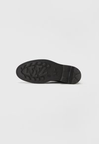 Semelle de chaussure en caoutchouc noir présentant un motif de grip texturé, une forme arrondie et un détail de logo en relief. Bords latéraux lisses et talon renforcé.