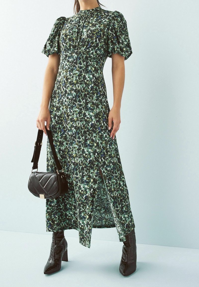 Femme portant une robe midi verte à fleurs avec manches bouffantes et fente devant, tenant un sac à main noir, associée à des bottines noires à talons.