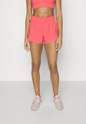 Vrouw draagt felroze sportshorts, bijpassende sportbeha en lichtroze sportschoenen, staand tegen een effen achtergrond.