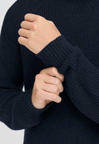 Temno mornarsko modra pletena jopica z rebrastim vzorcem. Izgled iz blizu, kjer roke vlečejo za rokavi, prikazuje zaključke in podrobnosti tkanine.