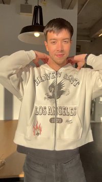 Ung mand justerer sin hoodie med ørn og "Heaven Sent Los Angeles 1971" tryk, stående indendørs under en hængelampe i sollys.