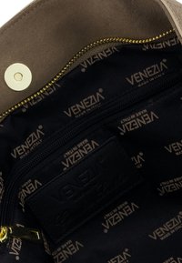 Wnętrze brązowej torby z zamszu z złotym zamkiem błyskawicznym, wykończone czarną tkaniną z wytłoczonym napisem "VENEZIA" oraz skórzaną metką.