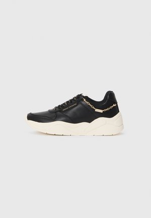 Black leather sneaker featuring a chunky cream sole, gold-tone chain accent, and black laces. Smooth texture with embossed branding on the side.