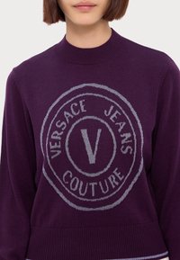 Pull violet avec un logo rond arborant l'inscription "VERSACE JEANS COUTURE" en lettres argentées. Col et ourlet côtelés avec une texture tricotée douce.