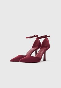 Burgundi spalvos aukštakulniai, smailiapirštiai, su kulkšnies dirželiais ir stiletto kulnais, ant balto fono.