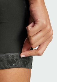 Black athletic shorts feature a textured, rubberized waistband with a logo pattern. A hand is pulling the waistband's edge for adjustment.