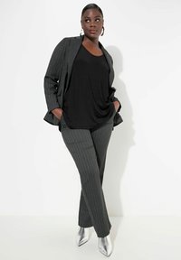 Musta drape-kotelo harmaan raidallisen bleiserin ja vastaavien housujen alla, yhdistettynä hopeisiin kärkitossuihin, neutraalilla taustalla.